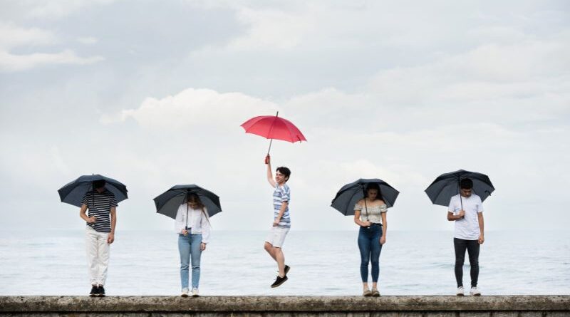A young man jumps joyfully holding a vibrant red umbrella while surrounded by four somber people holding black umbrellas against a cloudy coastal backdrop, symbolizing the breakthrough of existential depression.