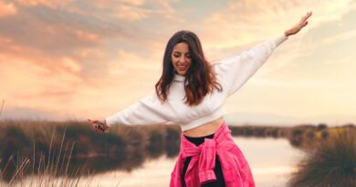 Een lachende vrouw staat in balans met gespreide armen op een muurtje bij een rivier tijdens zonsondergang, symboliserend het stoppen met jezelf wegcijferen en het herpakken van eigen kracht.