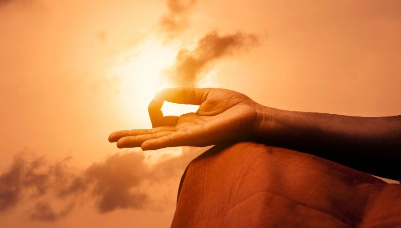Close-up of a hand in Gyan Mudra during meditation against a sunset sky, representing the fundamentals of yoga and spiritual union.