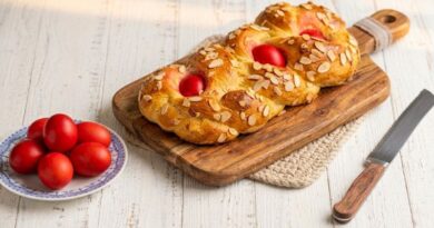 Een gevlochten Grieks paasbrood (Tsoureki) met amandelschaafsel en drie dieprood geverfde eieren op een rustieke houten broodplank met een broodmes ernaast.