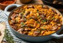 Close-up photo of a rustic blue enameled skillet filled with creamy Cajun mushroom pasta and tagliatelle on a wooden table. The dish is garnished with fresh parsley and surrounded by bowls of smoked paprika and herbs.
