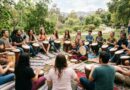 A diverse community group of people of all ages sitting in a large outdoor drum circle on blankets in a grassy park, actively playing djembe and other hand drums under natural daylight.