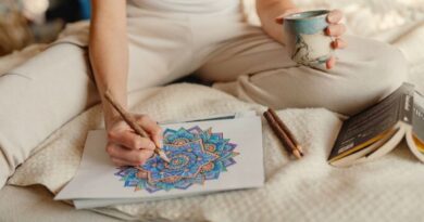 A person sitting comfortably, holding a warm drink while coloring an intricate geometric mandala with colored pencils for stress relief and relaxation.