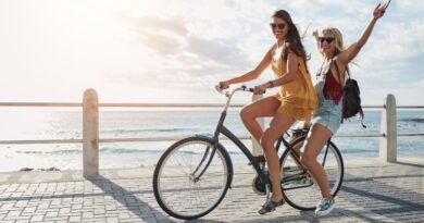 Twee lachende vriendinnen en geestverwanten op één fiets langs een zonnige boulevard aan zee, symbool voor de makkelijke klik tussen gelijkgestemden.