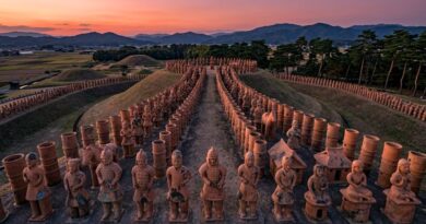 Een luchtfoto van een historisch Japanse Kofun-grafcomplex bij zonsondergang, geflankeerd door rijen terracotta Haniwa-figuren op een aarden pad. Op de achtergrond zijn grafheuvels, landbouwvelden en bergen zichtbaar.