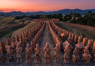Een luchtfoto van een historisch Japanse Kofun-grafcomplex bij zonsondergang, geflankeerd door rijen terracotta Haniwa-figuren op een aarden pad. Op de achtergrond zijn grafheuvels, landbouwvelden en bergen zichtbaar.