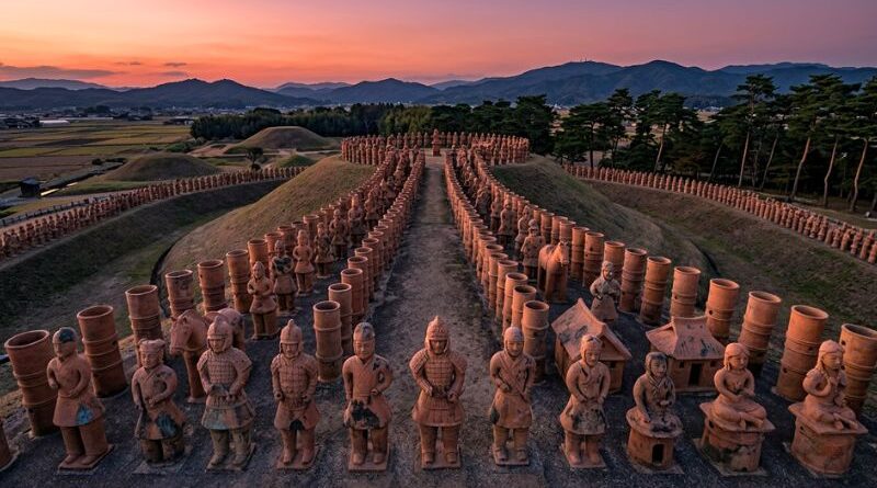 Een luchtfoto van een historisch Japanse Kofun-grafcomplex bij zonsondergang, geflankeerd door rijen terracotta Haniwa-figuren op een aarden pad. Op de achtergrond zijn grafheuvels, landbouwvelden en bergen zichtbaar.