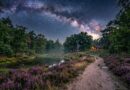 Sfeervolle nachtelijke landschapsfoto van een kronkelend zandpad door een bloeiend paars heideveld met een verlicht houten huisje onder de Melkweg