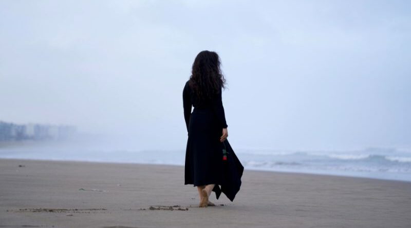 A woman with long curly hair walking barefoot on a quiet, misty beach while holding a single red rose, symbolizing self-empathy and softening the inner critic.
