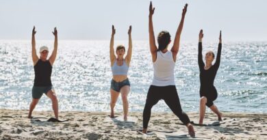 A small group of women practicing a yoga flow together on a sunny sandy beach facing a sparkling ocean, representing the science of daylife and benefits of daytime socializing.