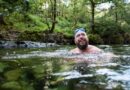 A smiling, bearded man enjoying the mental health benefits of cold water swimming in a clear, natural river surrounded by lush green trees.