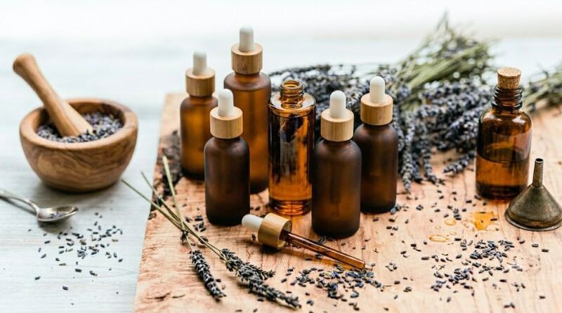 A rustic wooden table with amber glass dropper bottles filled with homemade lavender infused oil, surrounded by dried lavender flowers, a mortar and pestle, and a small funnel.