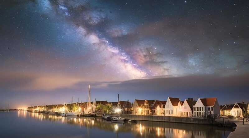 Een adembenemend nachtlandschap met de heldere Melkweg over een verlicht historisch vissersdorp en jachthaven bij rustig water.