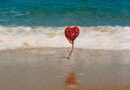 Een decoratief rood hart op een stokje dat in het zand aan de vloedlijn staat, met golvend zeewater op de achtergrond. Symboliseert de stabiele basis en liefde binnen een gezonde relatie.