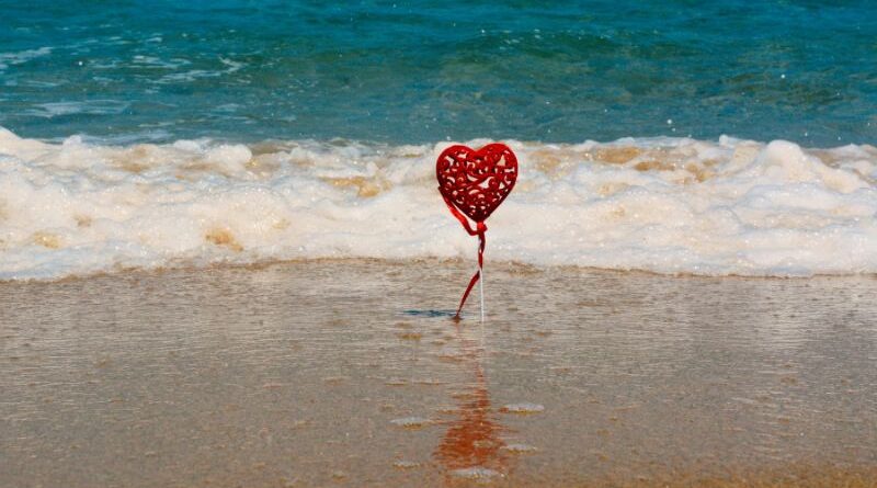 Een decoratief rood hart op een stokje dat in het zand aan de vloedlijn staat, met golvend zeewater op de achtergrond. Symboliseert de stabiele basis en liefde binnen een gezonde relatie.