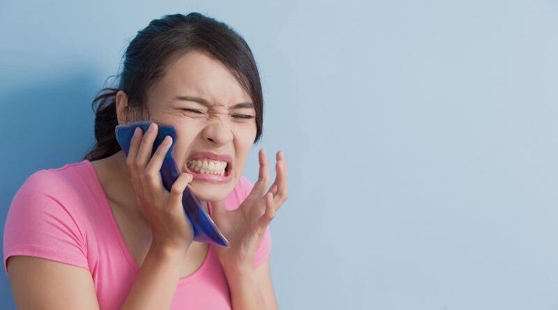 Een jonge vrouw in een roze shirt drukt pijnlijk kijkend een blauw koelkompres tegen haar rechterkaak vanwege hinderlijke mondpijn of tandpijn.