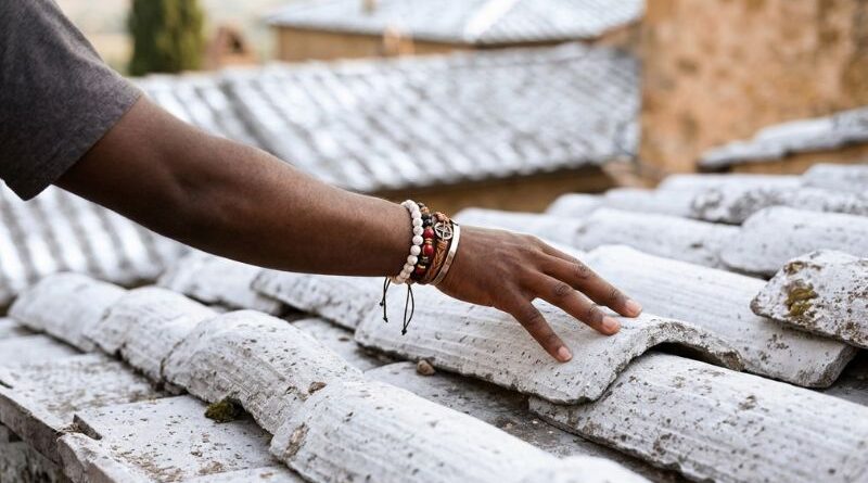 Close-up of a person's hand wearing beaded bracelets gently touching a freshly painted, white reflective tile roof to feel the cooling effect in an African township.