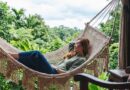 A smiling woman rests peacefully in a natural macrame fringe hammock on a jungle-view veranda, embodying the Vedic living philosophy at a tropical retreat. She is surrounded by dark wood furniture and a dense, lush rainforest canopy.