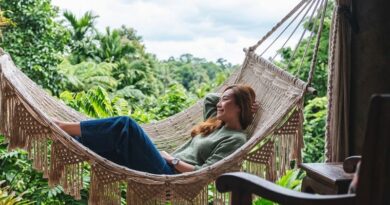 A smiling woman rests peacefully in a natural macrame fringe hammock on a jungle-view veranda, embodying the Vedic living philosophy at a tropical retreat. She is surrounded by dark wood furniture and a dense, lush rainforest canopy.