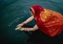 High-angle view of a woman in a traditional red and orange sari offering burning incense over dark water during a sacred ritual.