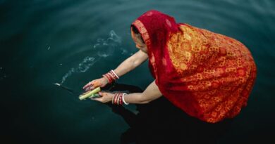 High-angle view of a woman in a traditional red and orange sari offering burning incense over dark water during a sacred ritual.