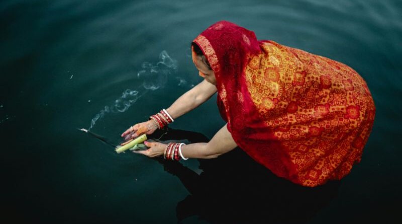 High-angle view of a woman in a traditional red and orange sari offering burning incense over dark water during a sacred ritual.