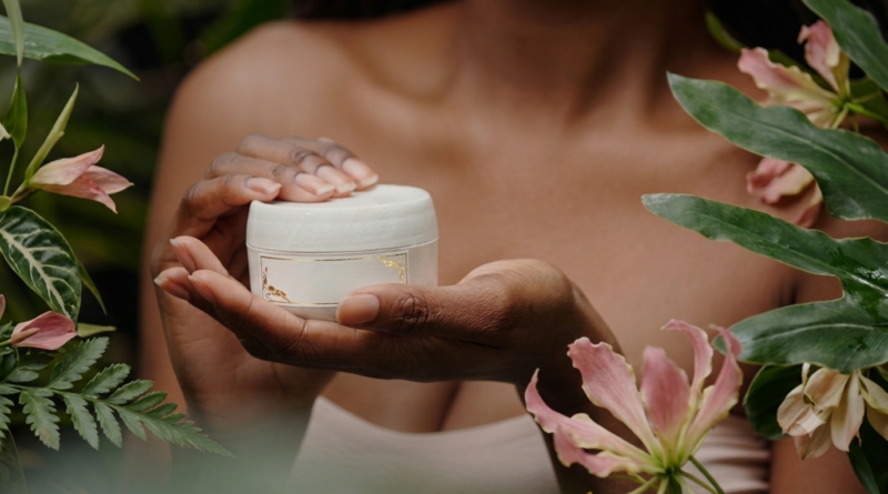 A close-up of a person of color's hands holding a white skincare jar with gold accents, framed by tropical greenery and pink flowers, representing Global Heart's natural beauty products.
