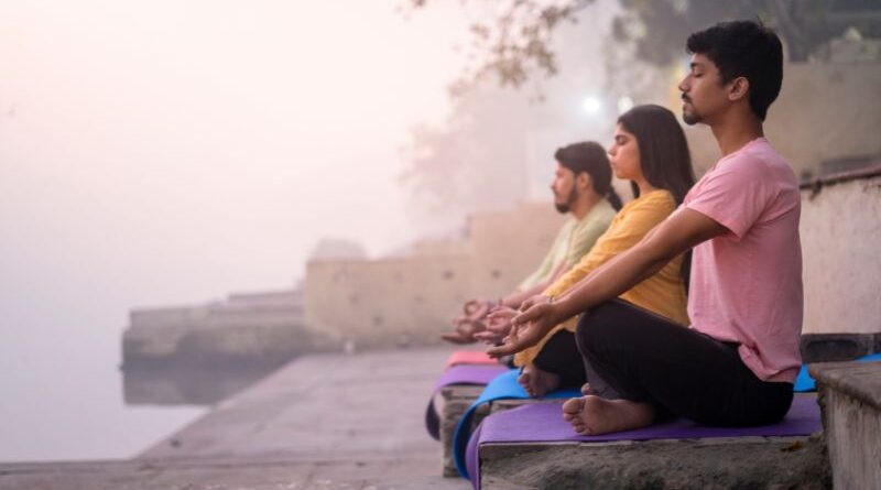 Drie mensen beoefenen Kapalbhati Pranayama vuurademhaling op yogamatten tijdens zonsopgang aan de oever van de Ganges rivier in India.