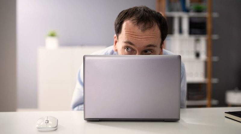 Een man op kantoor kijkt angstig en behoedzaam achter zijn laptop vandaan om zijn eigen energie te beschermen tegen energiezuigers op de werkvloer.